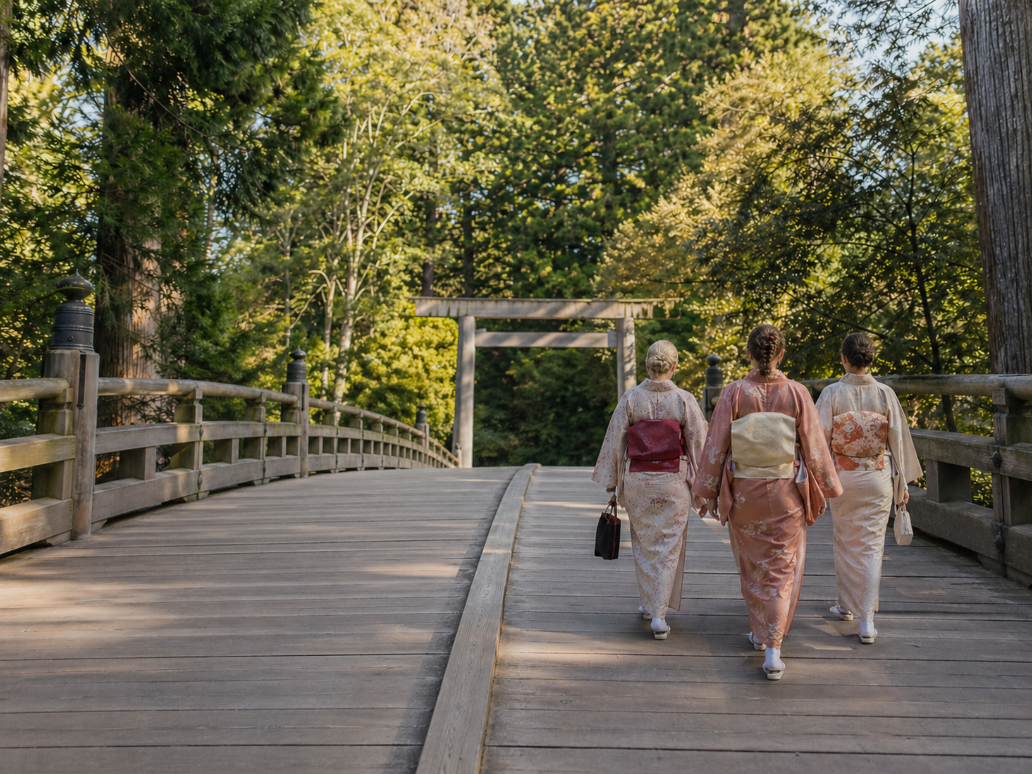 Three visitors in kimono-style attire walking across a wooden bridge toward a torii surrounded by forest